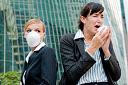 Picture of business woman sneezing, while another woman wearing a surgical mask looks on.